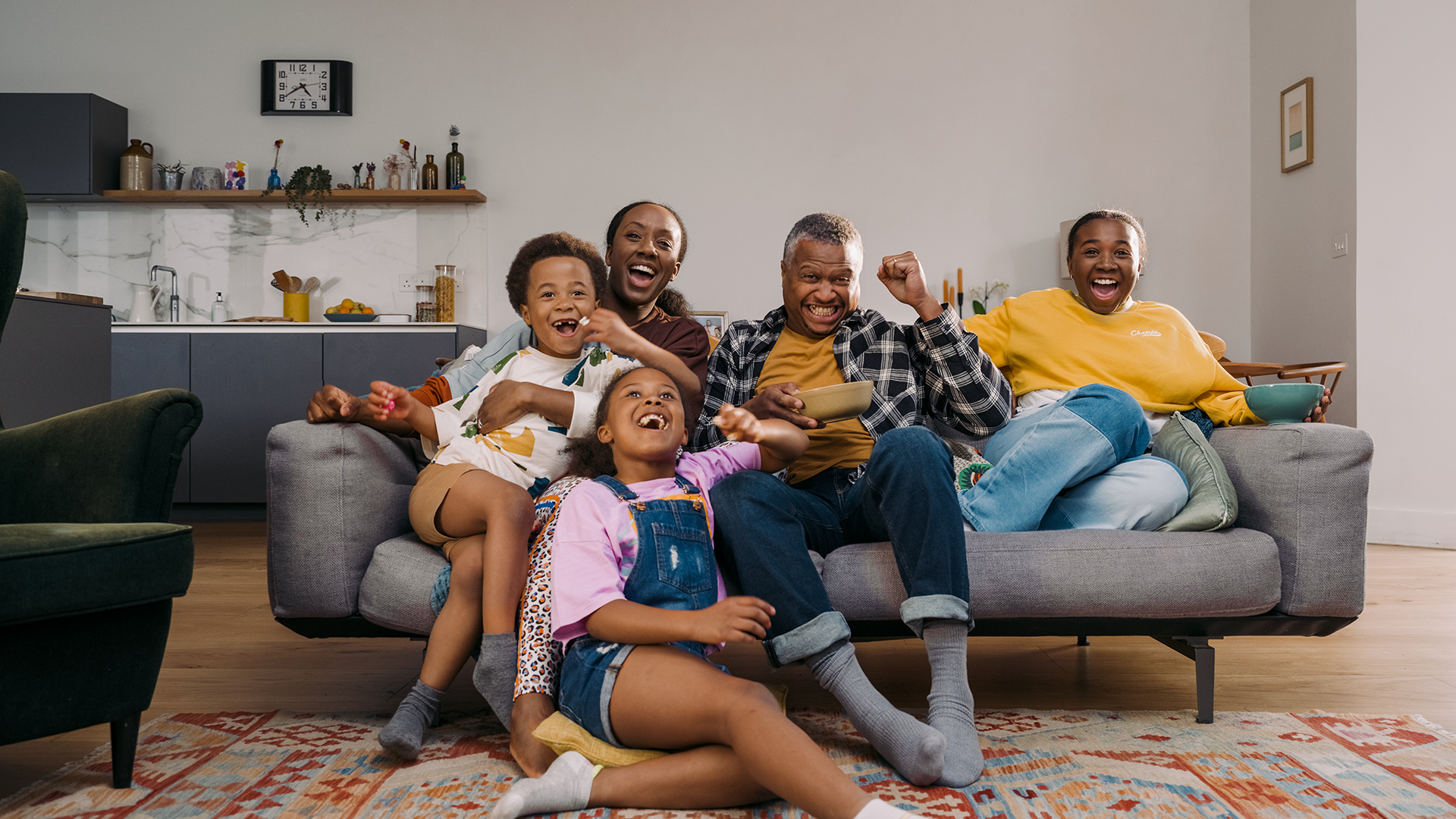 Family of five, including two parents and three young children, sat together on a sofa celebrating while watching television.