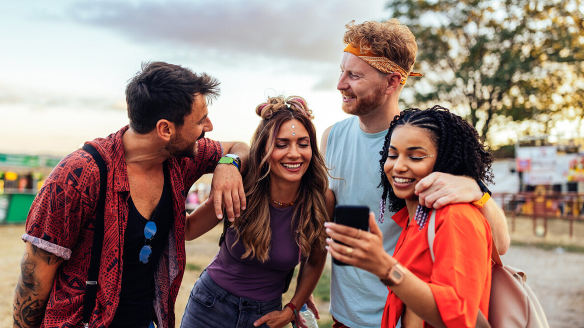 Group of young people smiling while huddled around a smartphone at a music festival.