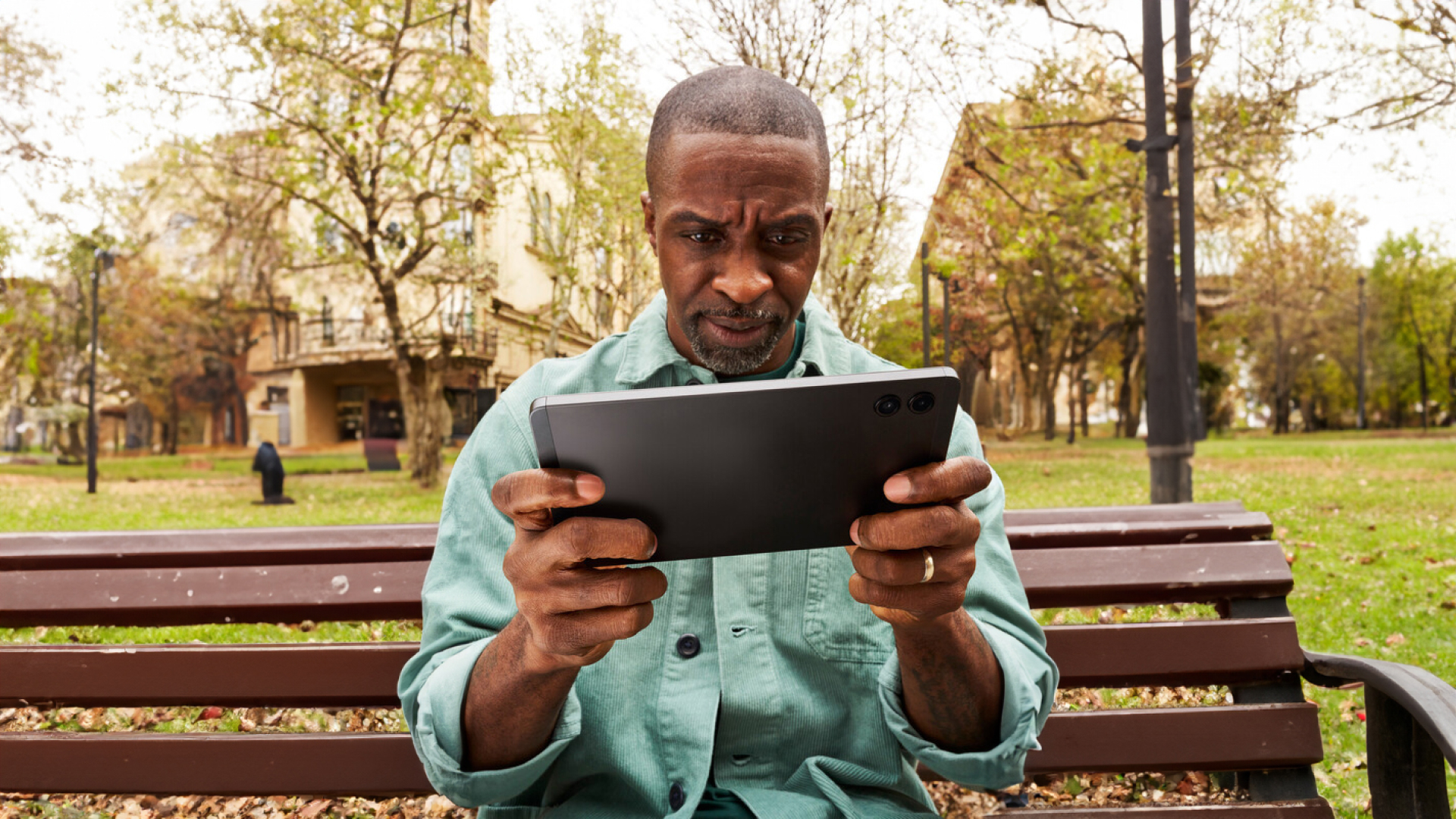 Man in aqua shirt streaming on his tablet on a park bench with trees in the background.