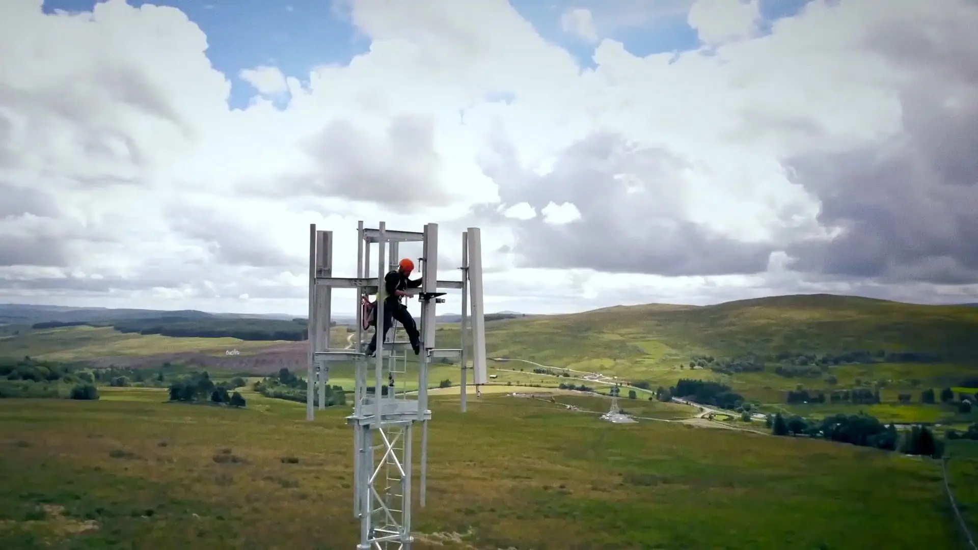 An Openreach engineer works high up on a telecommunications mast, overlooking green fields.