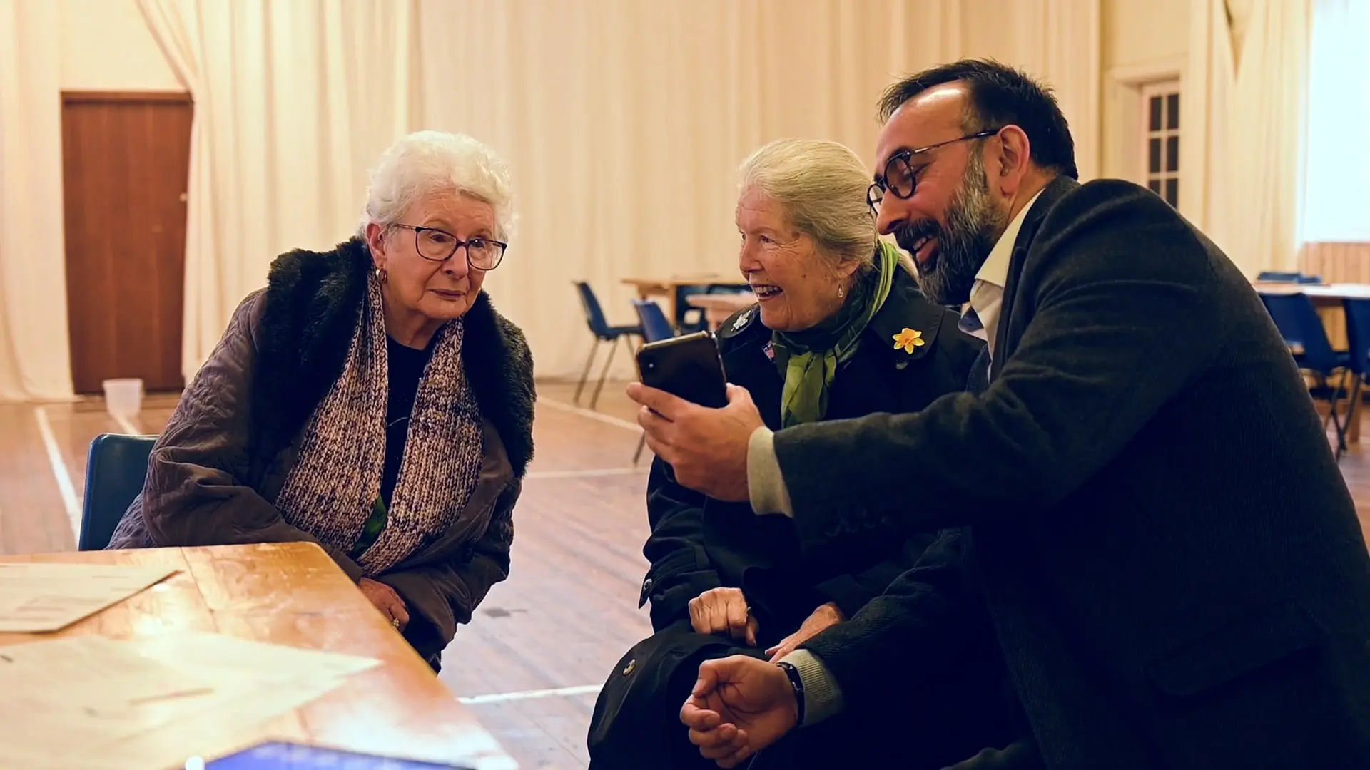 A man demonstrates a task on a mobile phone, while two senior women watch.