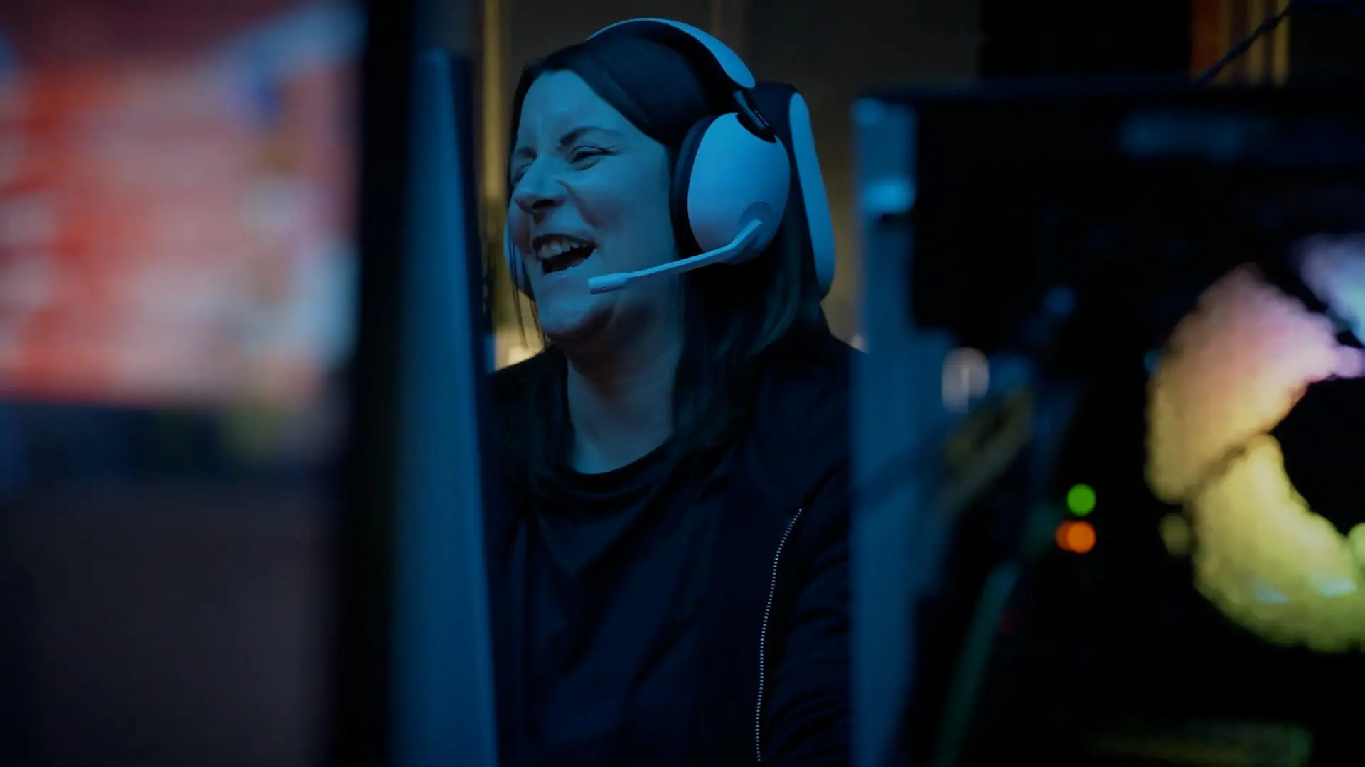 Through the gap between two gaming computers with bright lights, a woman in a gaming headset is laughing.