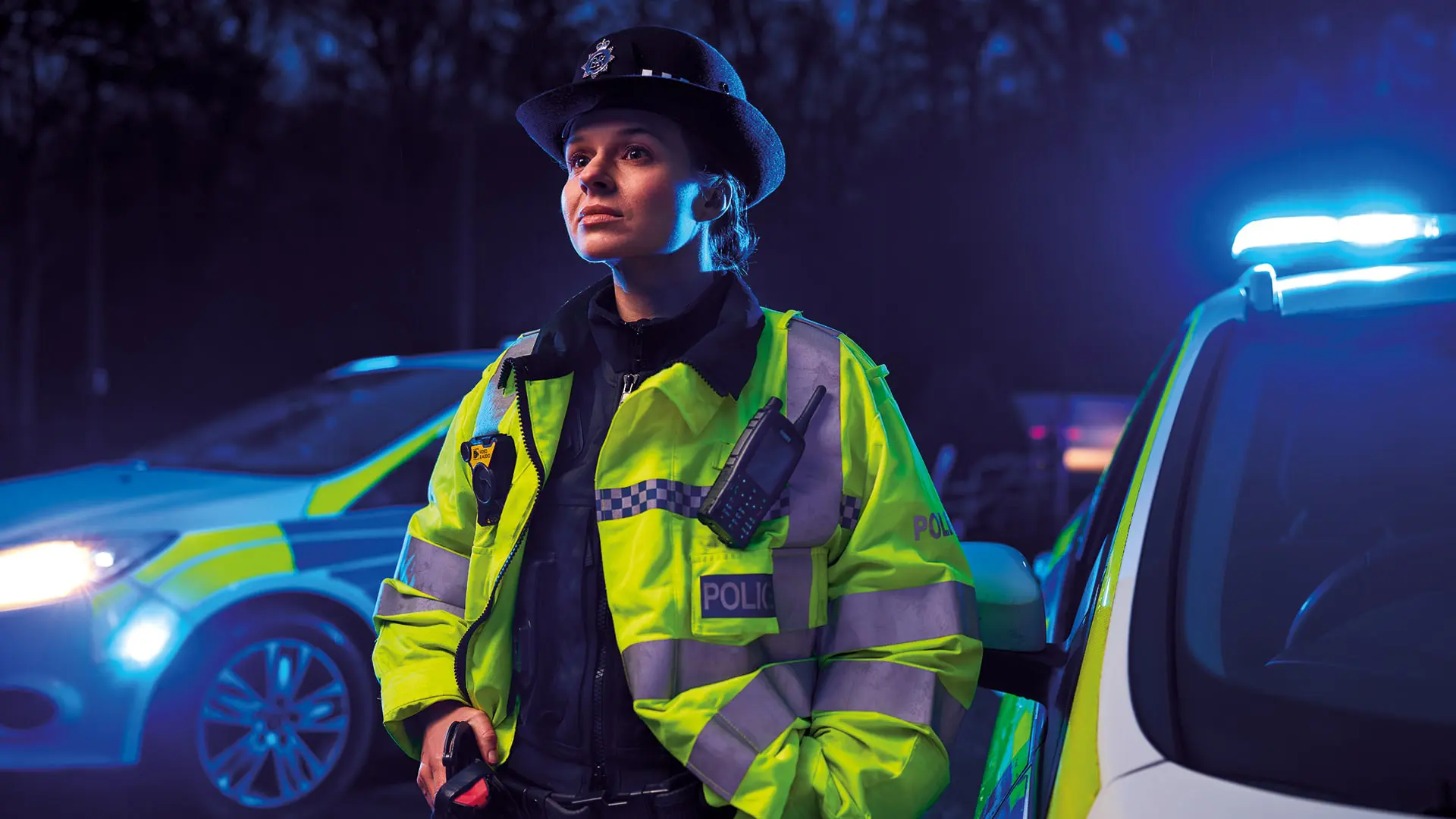 A policewoman stands in front of a police car, lights flashing, at night.